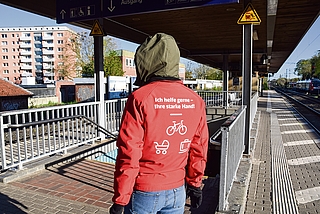 Am Elmshorner Bahnhof steht ein Reisendenhelfer auf dem Bahnsteig zwischen den Gleisen 2 und 3. Er bietet Fahrgästen mit schweren Koffern, Kinderwagen und E-Bikes seine Hilfe an. Im Hintergrund sind die Bretterwände zu sehen, mit denen der Fahrstuhlschacht abgesperrt ist – voraussichtlich noch bis Anfang September. (Foto: Frank)