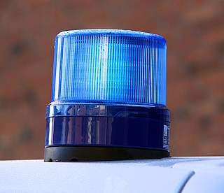 Eine Nahaufnahme einer blauen Warnblinkleuchte, die an einem Fahrzeug angebracht ist, vor einem unscharfen braunen Hintergrund. Die Leuchte ist eingeschaltet und leuchtet hell.