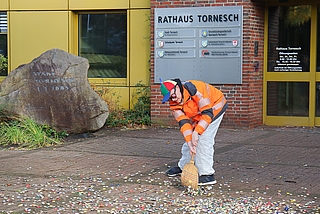 Ausgerüstet mit einem Reisigbesen, einer Warnweste und einer Brille mit runden „30“-Schildern fegt Daniel Kölbl vor dem Tornescher Rathaus Kronkorken zusammen. (Foto: CDU Tornesch)