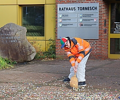 Ausgerüstet mit einem Reisigbesen, einer Warnweste und einer Brille mit runden „30“-Schildern fegt Daniel Kölbl vor dem Tornescher Rathaus Kronkorken zusammen. (Foto: CDU Tornesch)