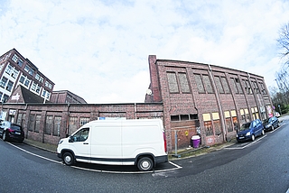 Dieses historische Ensemble an der Schloßstraße steht unter Denkmalschutz: Das zweistöckige Citastrom-Haus (rechts), die niedrige Mantelhalle (in der Mitte hinter dem Lieferwagen) und das Kranhaus mit dem kleinen Giebel (links). Das Foto entstand mit einem Weitwinkelobjektiv. (Foto: Schröckert)