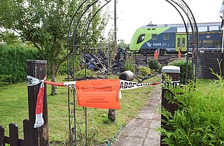 Die Polizei hat den Brandort in der Kleingartenanlage Fuchsberg in Elmshorn abgesperrt. Ein Regionalzug fährt im Schritttempo daran vorbei. Mitarbeiter der Deutschen Bahn untersuchen die technischen Anlagen. (Foto: Frank)