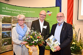 Vorstand Axel Huckfeldt (Mitte) verabschiedete John-Hermann Cordes (rechts), der 45 Jahre ehrenamtlich im Baumschulverband tätig war. Cordes engagierte sich im Juniorenkreis der Baumschuler, seit 1990 im Fachausschuss Obstgehölze auf Landesebene, seit 2005 bis 2023 als Vorsitzender des Obst-Fachausschusses und als Mitglied des Erweiterten Hauptausschusses auf Bundesebene. In den 45 Jahren hielt ihm seine Frau Petra (links) den Rücken frei und unterstützte das Engagement ihres Mannes. (Foto, M. Wienert, BdBSH)