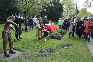 Fast 50 Menschen feierten in einer Regenpause die Einweihung der Gärtnerin-skulptur im Rosarium. Elsa Plath (Mitte vorn) trug die alte Ballade „Es waren zwei Königskinder“ vor. Sie spielte damit auf das Gegenüber der Figuren von Gärtner und Gärtnerin an. Der Künstler Ewald Rumpf (links) begleitete sie an der Violine. Viele Gäste stimmen in den Gesang ein. (Foto: Frank)